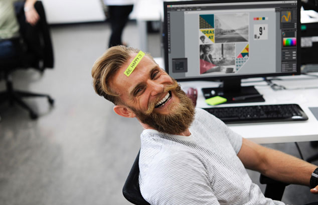 n homme portant une barbe de hipster et un post-it coll&eacute; sur le front disant &ldquo;Be Happy&rdquo; est assis &agrave; son bureau.