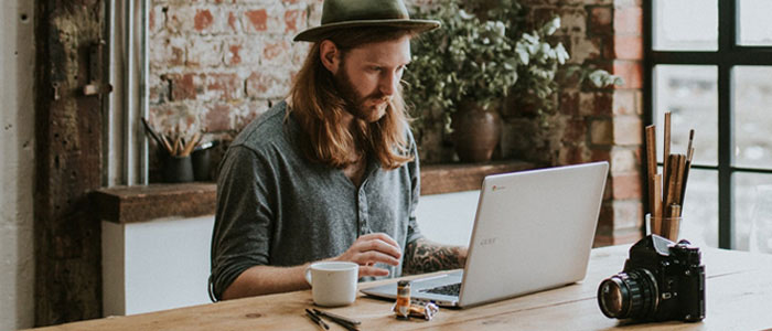 Jeune homme aux cheveux longs et &agrave; la barbe pleine assis dans un espace de travail regardant l&rsquo;ordinateur portable devant lui.