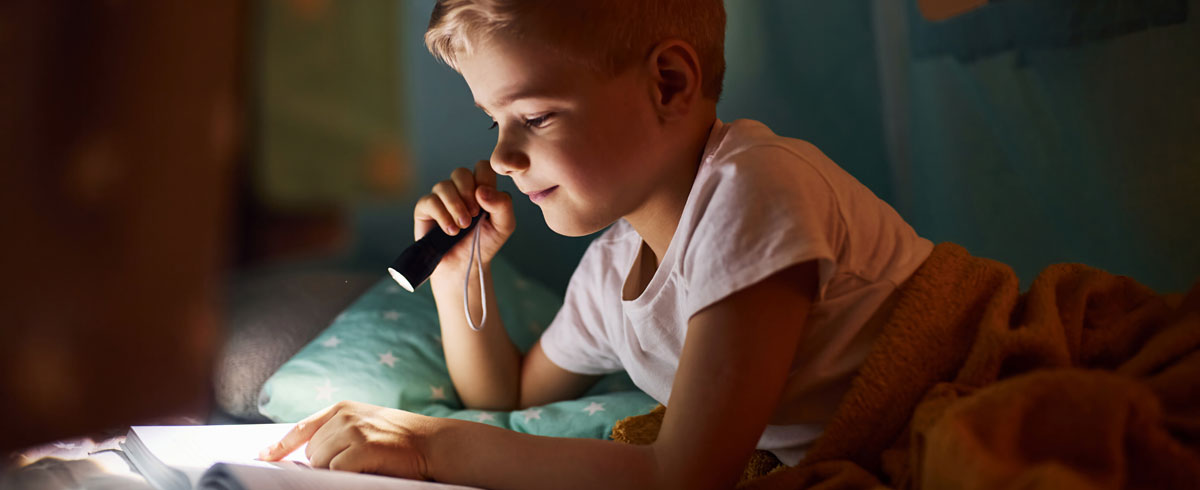 Un gar&ccedil;on lisant un livre avec une lampe torche &agrave; la main, allong&eacute; sur son lit avec une couverture