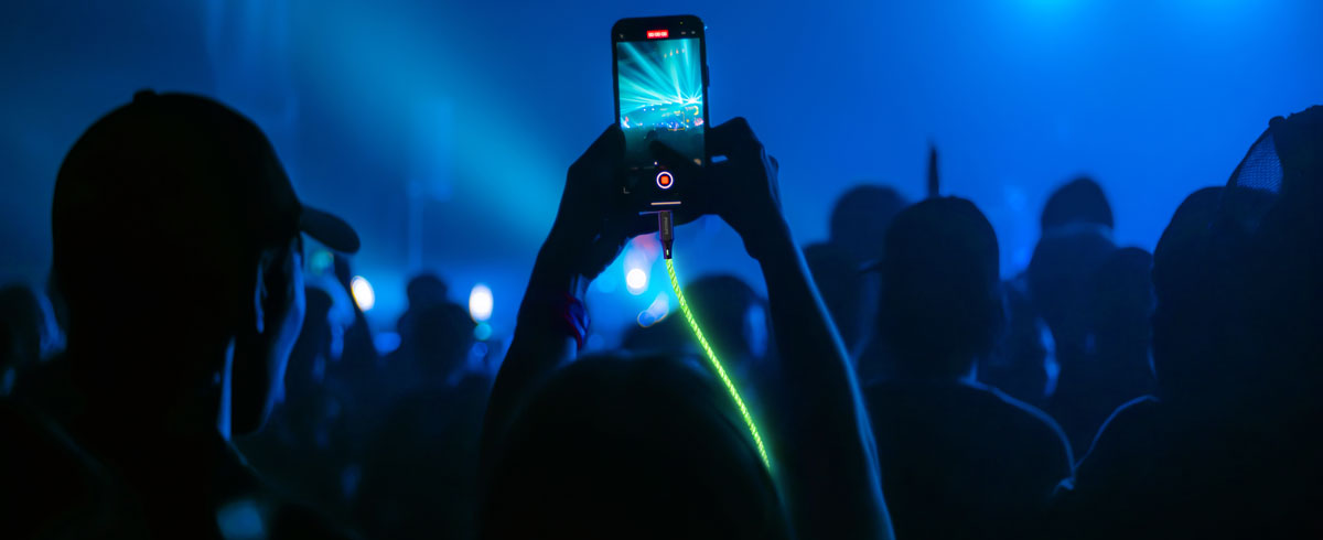 Une personne levant un t&eacute;l&eacute;phone portable connect&eacute; &agrave; un c&acirc;ble lumineux au milieu d'une foule, capturant l'instant pr&eacute;sent pendant un &eacute;v&eacute;nement